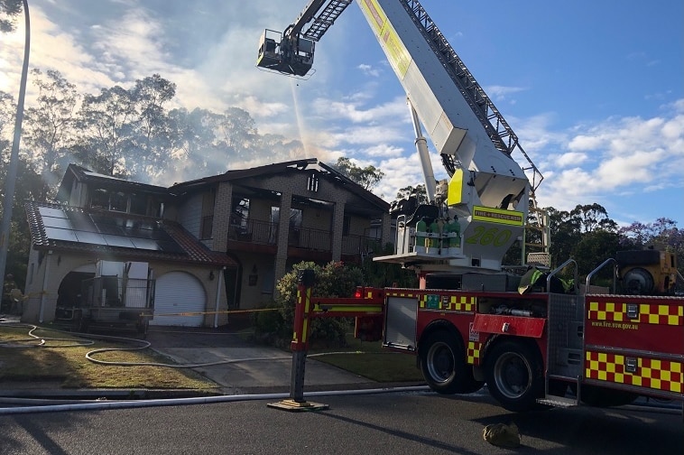 A fire truck with a long ladder extended over a two-storey house, shooting water into the fire-damaged roof of the house