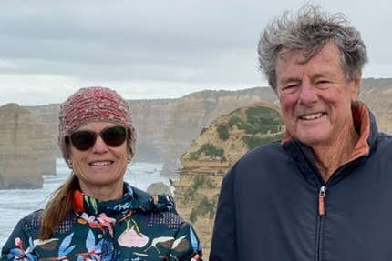 A man and a woman stand above a rugged coastline smiling at the camera.