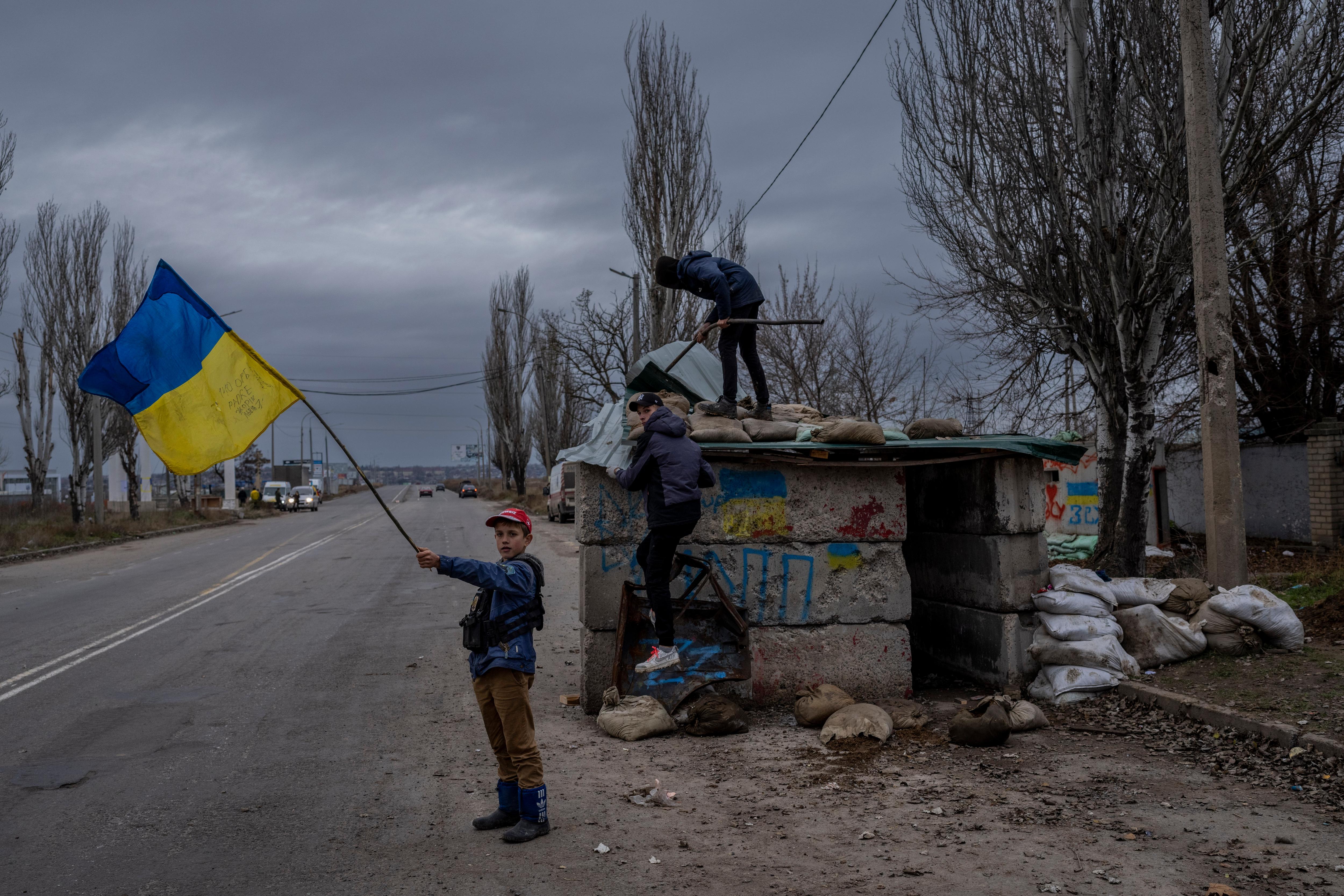Child waves large Ukrainian flag beside abandoned checkpoint as two others climb structure. 
