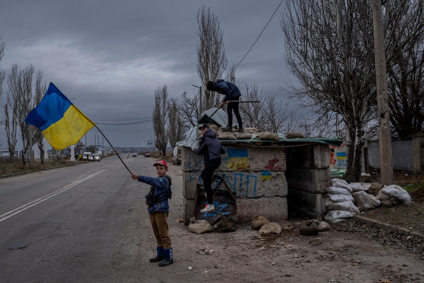 Child waves large Ukrainian flag beside abandoned checkpoint as two others climb structure. 