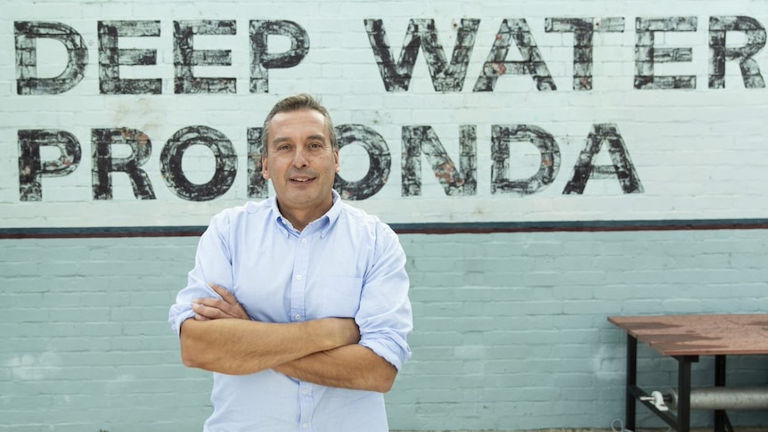 Christos Tsiolkas crosses his arms and stands at a pool in front of signage declaring "deep water".