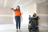 A man is a wheelchair inside a home yet to be painted next to a woman walking and pointing