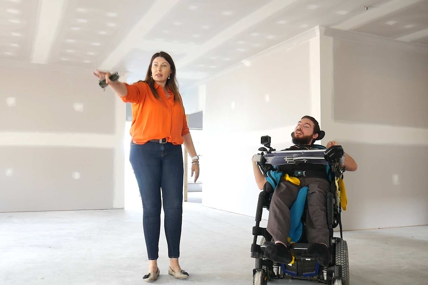 A man is a wheelchair inside a home yet to be painted next to a woman walking and pointing