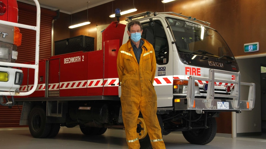 man wearing blue face mask in fire fighting outfit stands next to big red fire truck
