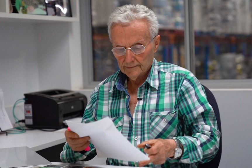 An older man in a check shirt holding a piece of paper which he is looking at. He is at a desk with printer in the background.