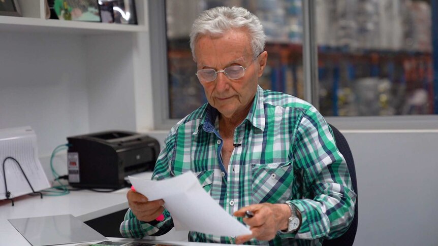 An older man in a check shirt holding a piece of paper which he is looking at. He is at a desk with printer in the background.