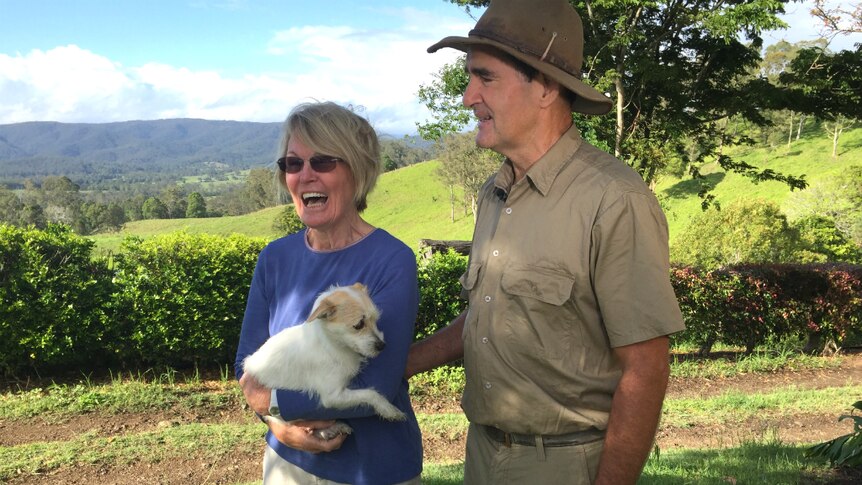 man and woman standing with a small white dog in the country, smiling and happy