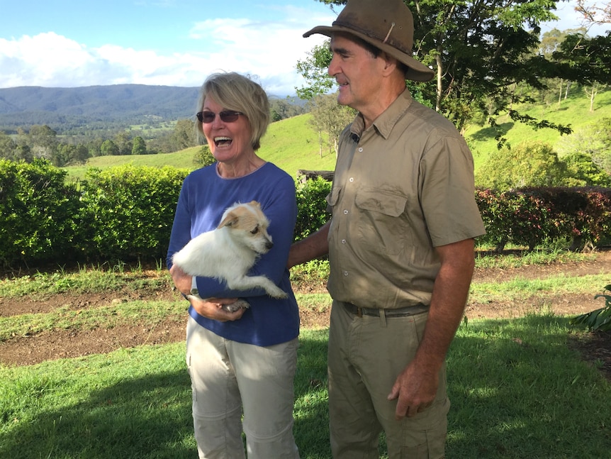 man and woman standing with a small white dog in the country, smiling and happy