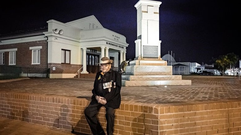 An older man in army hat with jacket and medals sits in front of a war memorial in early dawn light.