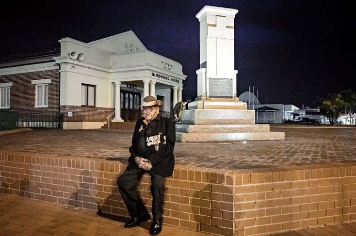 An older man in army hat with jacket and medals sits in front of a war memorial in early dawn light.