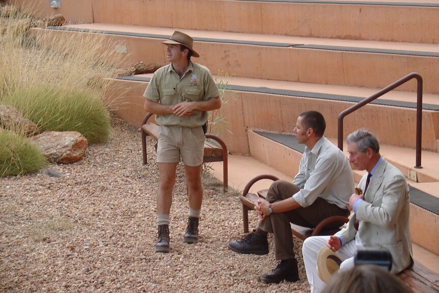 Prince Charles sits near another man as a third man in a khaki ranger-style uniform stands nearby.