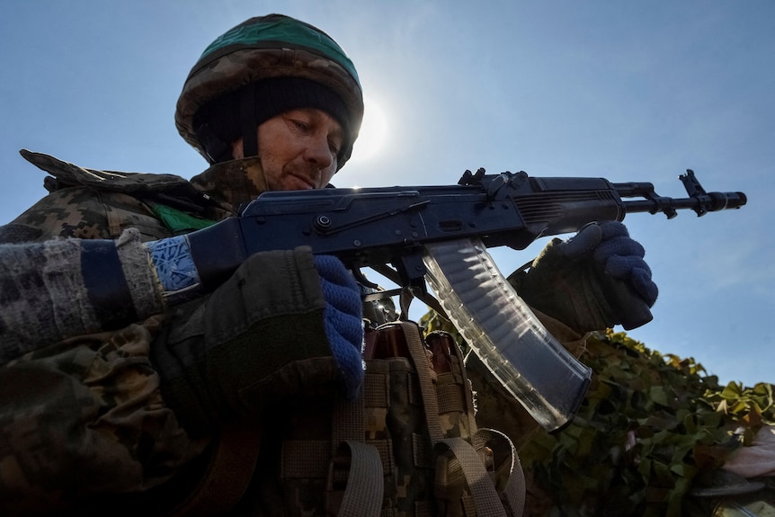A Ukrainian soldier wearing gloves and a helmet holds an AK-47 with bullets shown in a clear magazine.