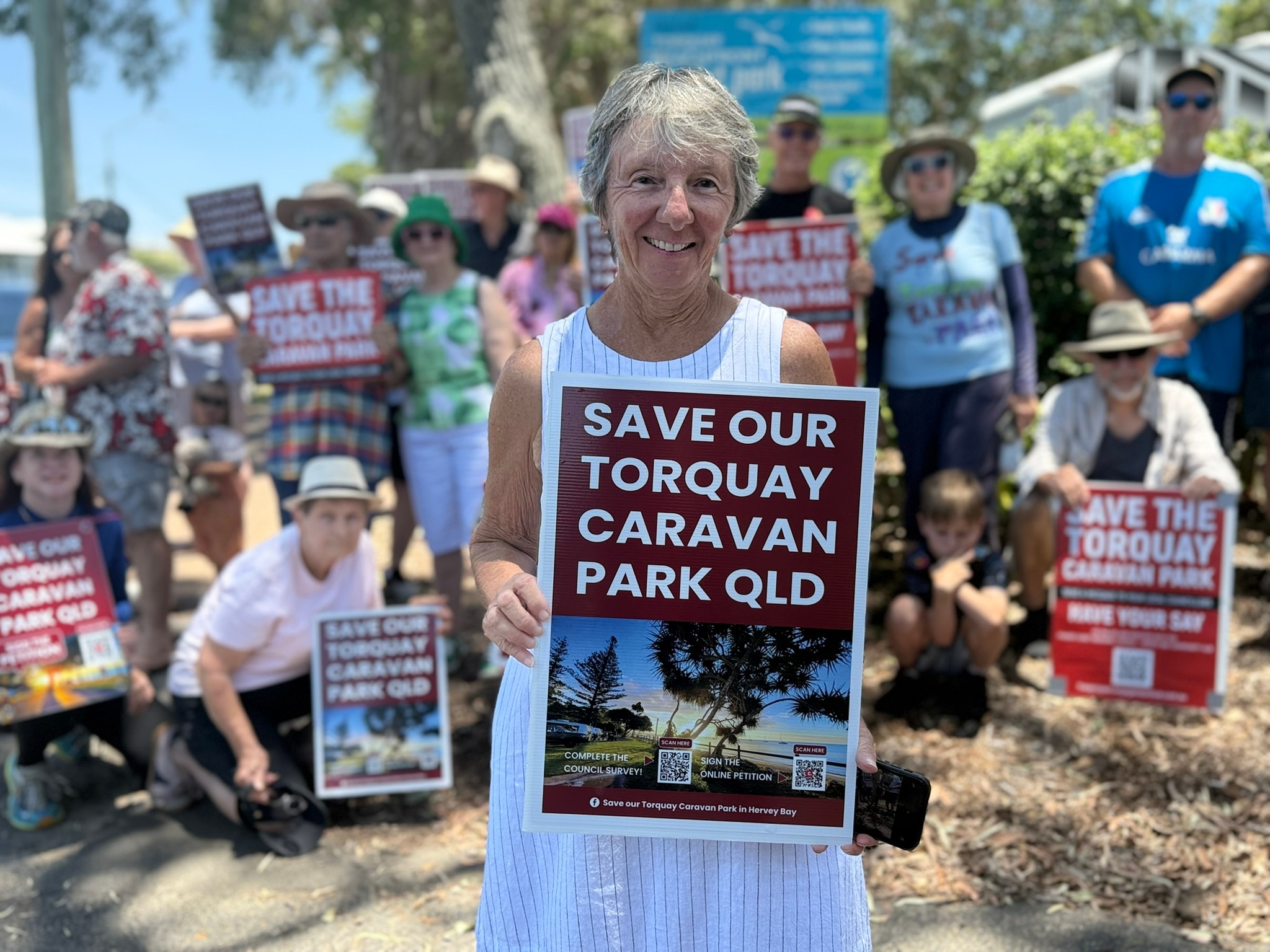 Uma mulher idosa sorridente segura uma placa diante de um grupo de pessoas que pede que um parque de caravanas permaneça aberto.