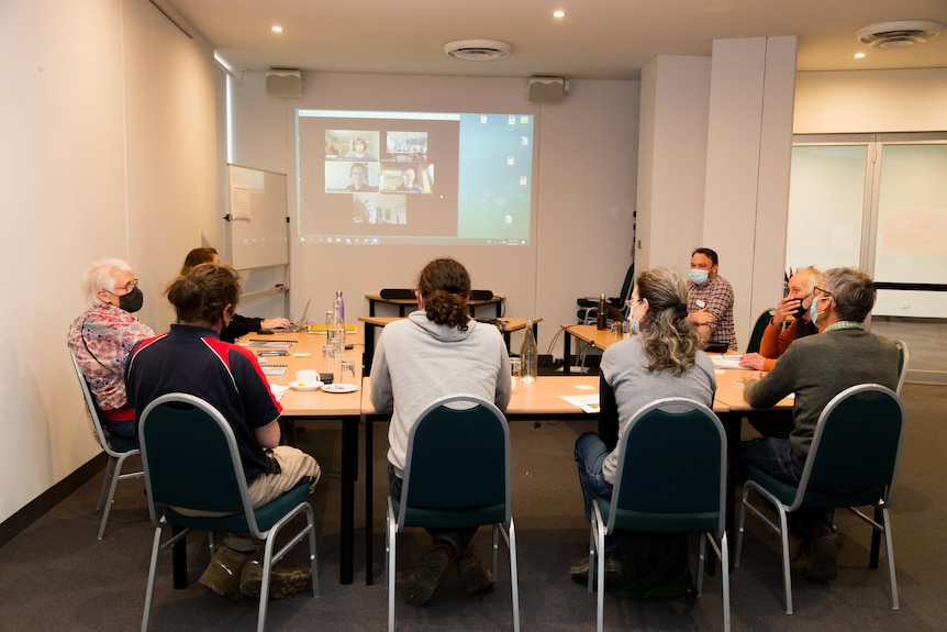Seven people sit in a meeting room, with more people on a screen on Zoom.