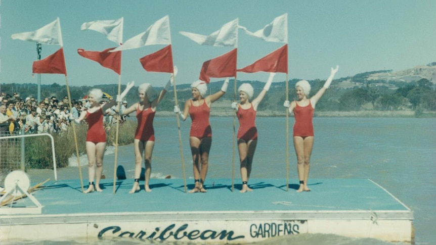 Women stand on a lake pontoon in swimwear holding flags.