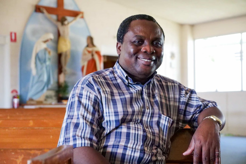 Father Daniel Djodjowuu Mawuko sits down in St Colomba's Church with the cross in the background.