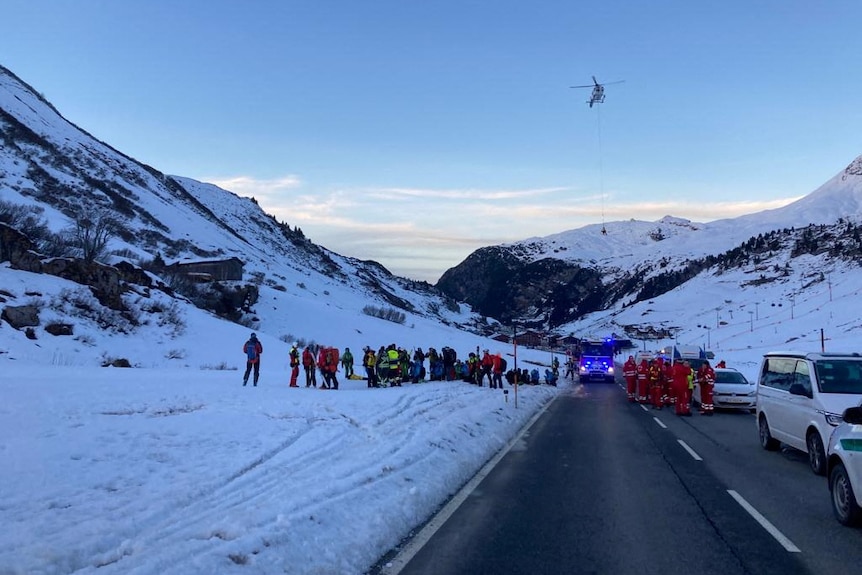 A road is pictured with cars and rescue workers and on the left a snowy field with a group of people. In the sky a helicopter.