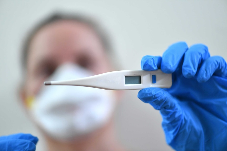 A person holds a thermometer up to the camera. The person is wearing a medical mask and their face is out of focus.
