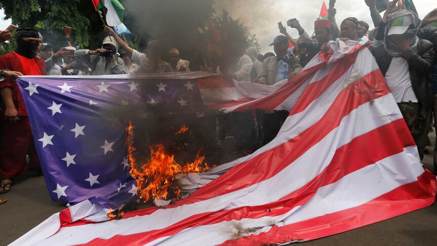 A group of protesters hold up a US flag as fire burns a hole in the middle of the flag.