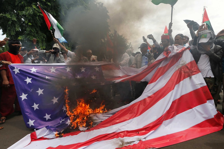 A group of protesters hold up a US flag as fire burns a hole in the middle of the flag.