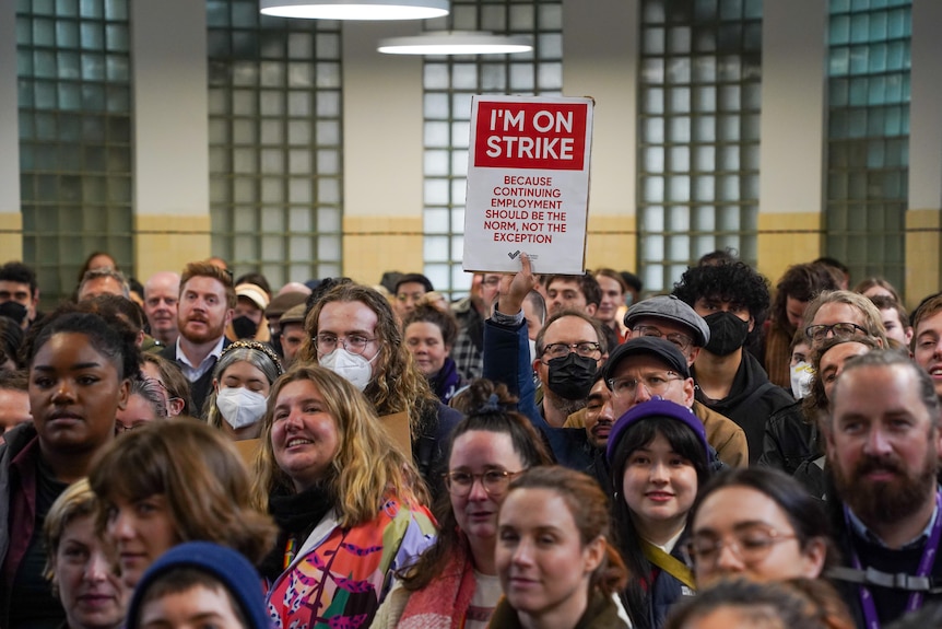 A crowd of dozens of university staff are seen filling Trades Hall, with someone holding up a protest sign near the back.