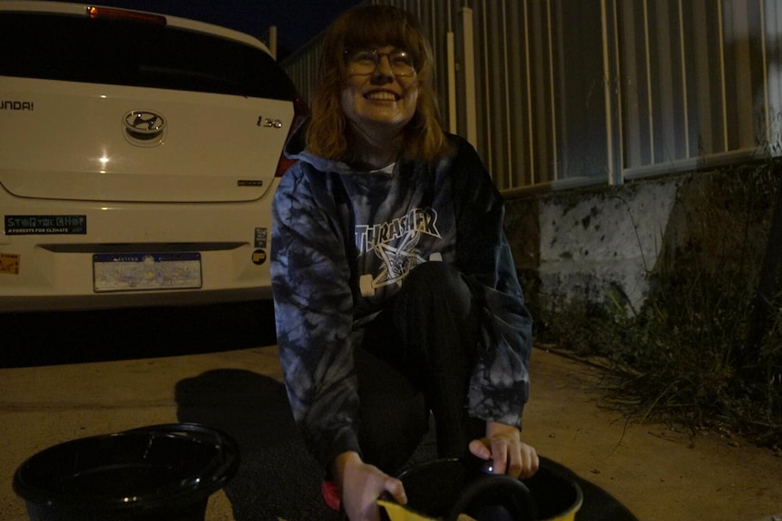 A young woman with red hair wearing a hoodie crouches in front of a car. 