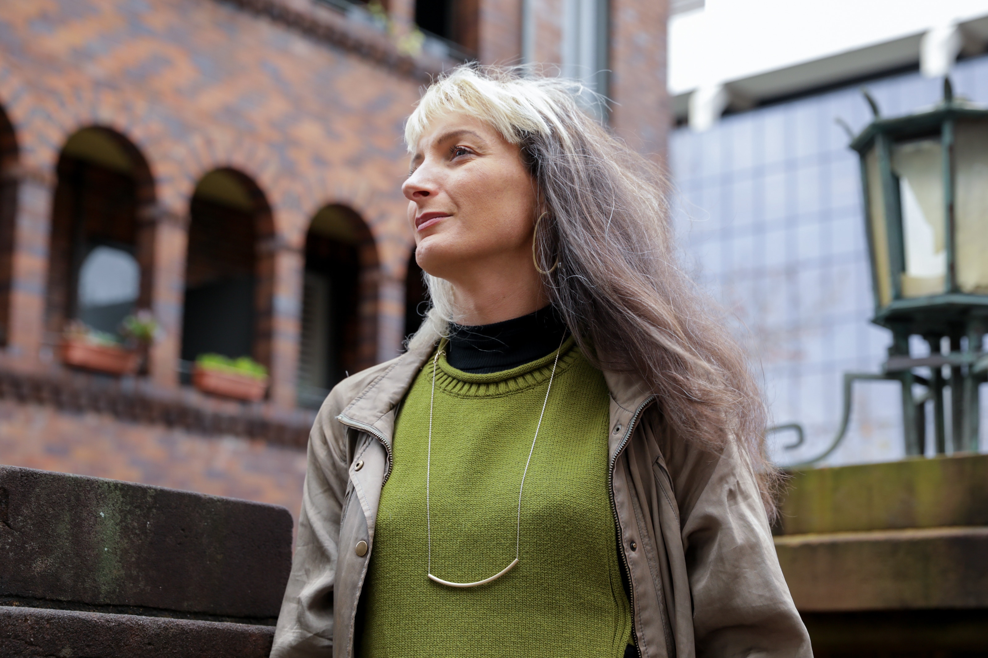 Una mujer blanca con cabello blanco y castaño está afuera, vestida con un suéter y un abrigo verdes, mirando directamente a la cámara.