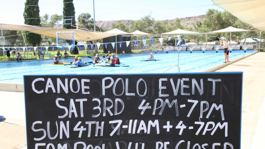 A sign for the canoe polo event in Alice Springs, while players train in the background.