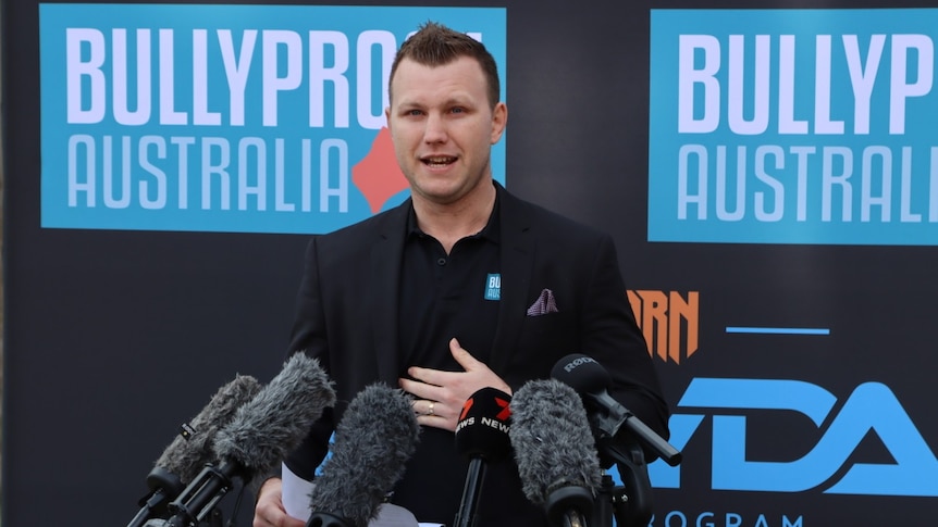 A man in a black t-shirt stands in front of 6 microphones