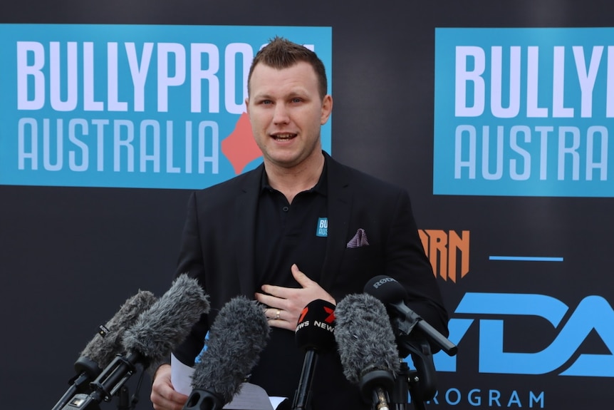 A man in a black t-shirt stands in front of 6 microphones