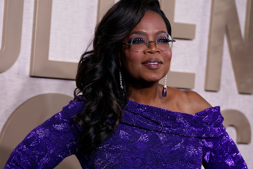 A close up shot of Oprah on a red carpet. She's wearing glasses and an off-the-shoulder purple dress.