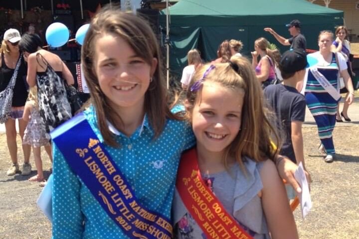 A young girl wearing a blue show sash stands next to another girl wearing a red show sash.