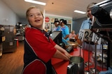A happy young boy behind the counter of a cafe, wears a red and blue t-shirt as a woman looks on.