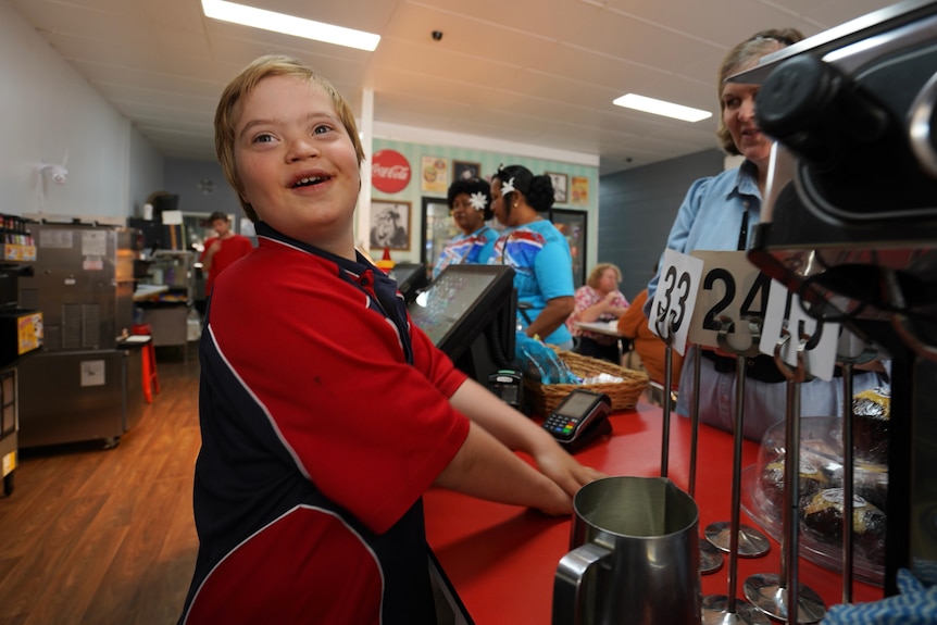 A happy young boy behind the counter of a cafe, wears a red and blue t-shirt as a woman looks on.