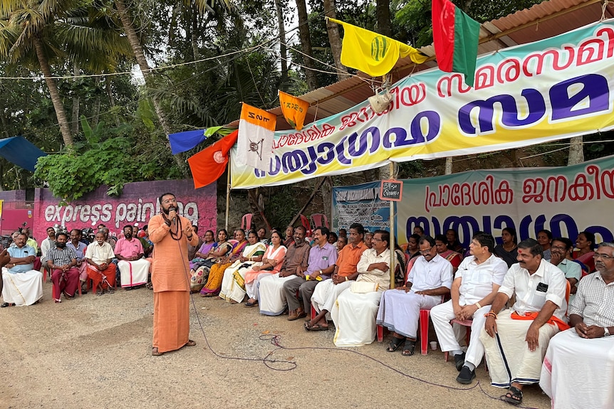 A man speaks into a microphone as people sit listening on chairs in rows next to him. 