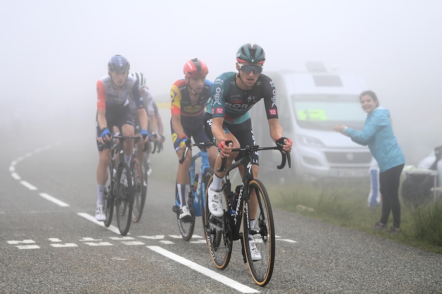 An Australian cyclist rides out of the fog ahead of two rivals, as a spectator applauds on the side of the road.