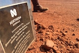 A sign explaining a new mine sits in red dirt as a person walks past in boots