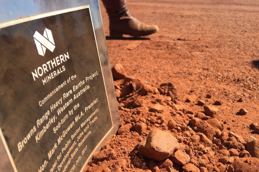 A sign explaining a new mine sits in red dirt as a person walks past in boots