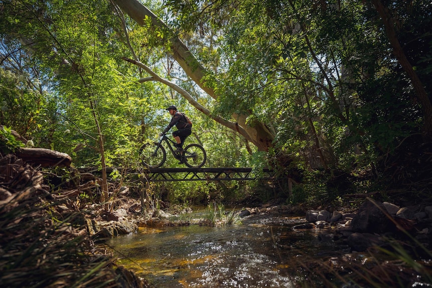 A man on a bike wearing a helmet goes over a bridge, with a river underneath. 