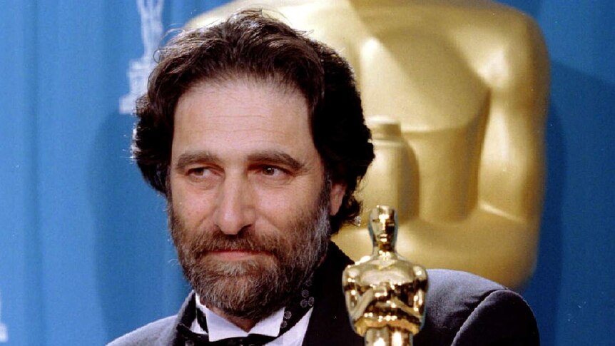 Eric Roth stands in a tuxedo holding an Oscar trophy