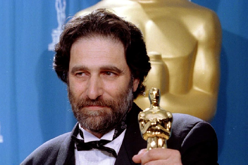 Eric Roth stands in a tuxedo holding an Oscar trophy