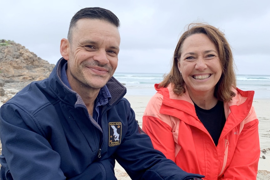 A woman with short brown hair wearing a bright pink coat sits on the shore with a man wearing a blue jacket, smiling.