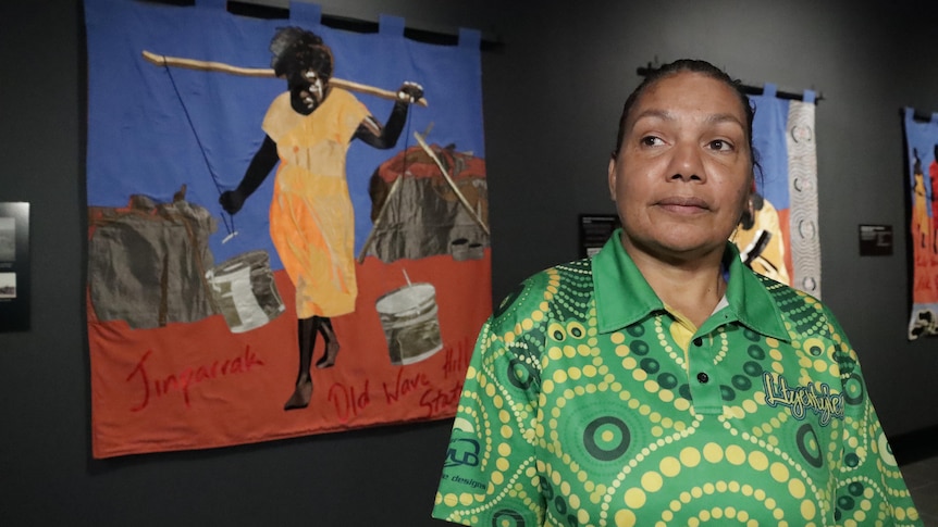 An Aboriginal woman stands in front of a textile mural depicting a woman carrying buckets of water