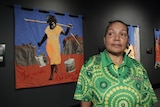 An Aboriginal woman stands in front of a textile mural depicting a woman carrying buckets of water