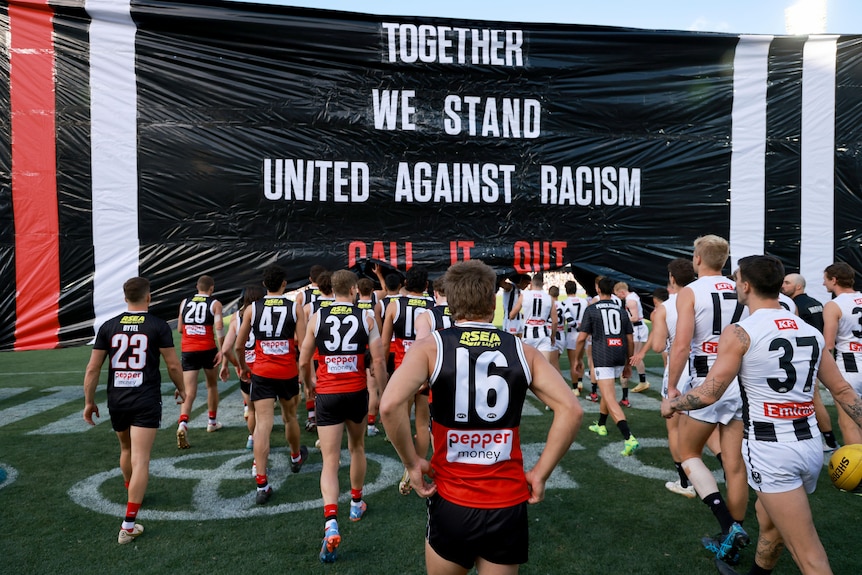 Collingwood and St Kilda players walk through a banner together which reads "TOGETHER WE STAND UNITED AGAINST RACISM"