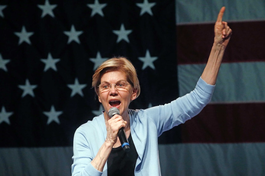 Elizabeth Warren wearing a powder blue cardigan, pointing one finger to the sky in front of a large US flag.
