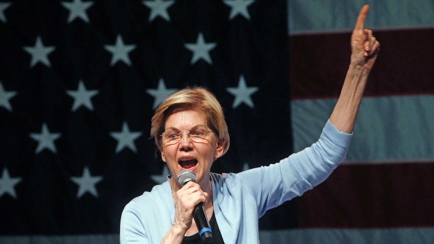 Elizabeth Warren wearing a powder blue cardigan, pointing one finger to the sky in front of a large US flag.