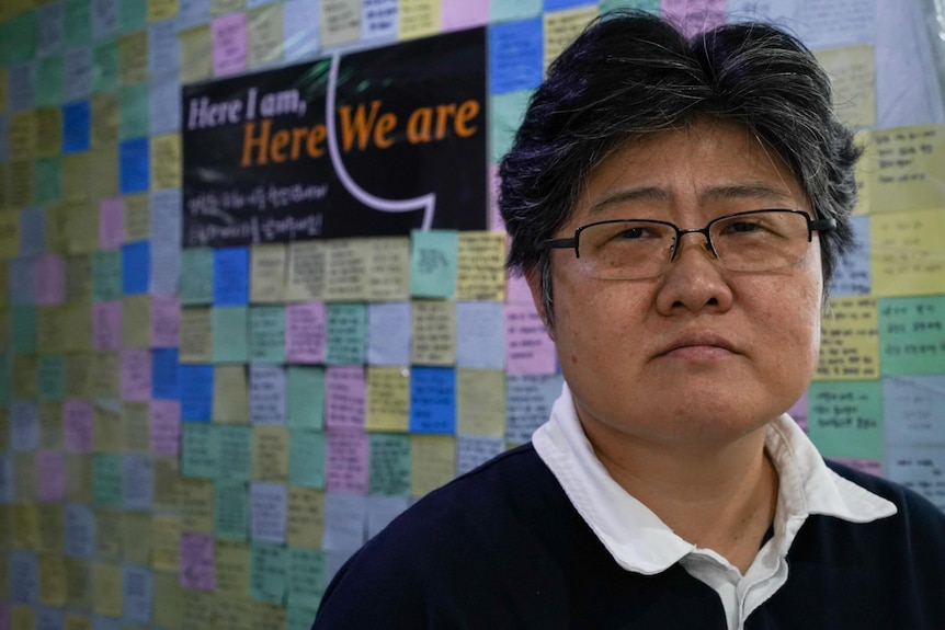 A close up of a woman with glasses in front of a poster covered in sticky notes