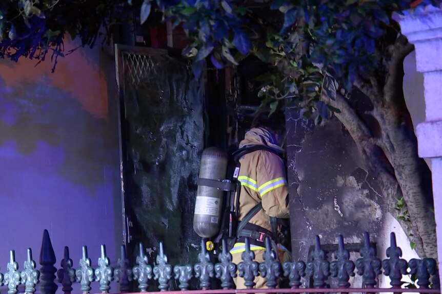 A firefighter walks into a burnt house with visible damage to the front at night.