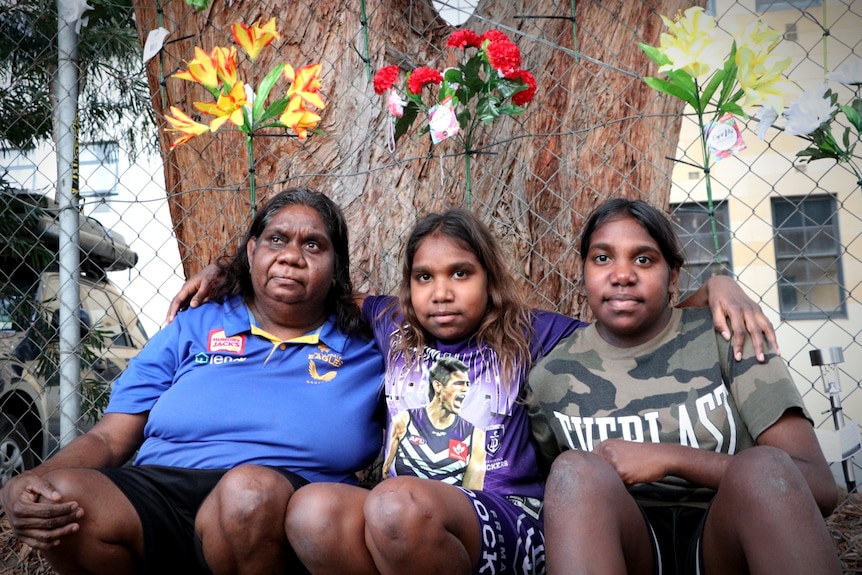 Three Aboriginal women sat in front of a wire fence with flower tributes pinned to it